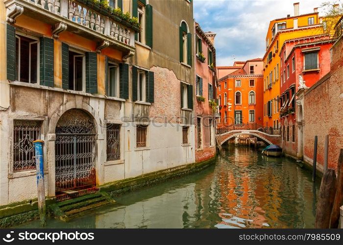 Colorful narrow lateral canal and pedestrian bridge in Venice with docked boats, Italy
