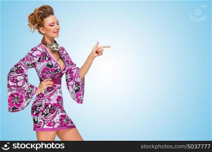 Colorful photo of a happy fashionable hippie homemaker with metal vintage music headphones around her neck, pointing aside with her finger on blue background.