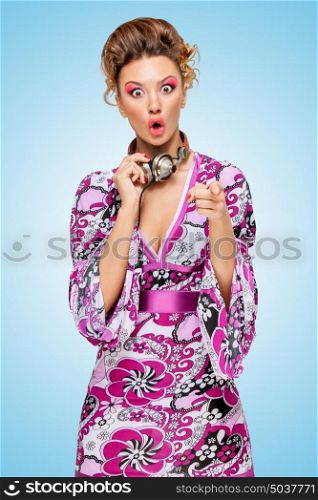 Colorful photo of an amazed fashionable hippie homemaker with metal vintage music headphones around her neck, pointing to the camera with her finger on blue background.