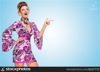 Colorful photo of an amazed fashionable hippie homemaker with metal vintage music headphones around her neck, pointing aside with her finger on blue background.