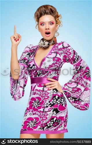 Colorful photo of an amazed fashionable hippie homemaker with metal vintage music headphones around her neck, pointing up with her finger on blue background.