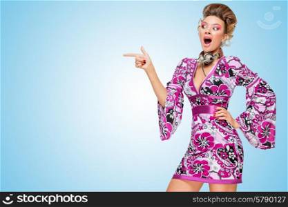 Colorful photo of an amazed fashionable hippie homemaker with metal vintage music headphones around her neck, pointing aside with her finger on blue background.