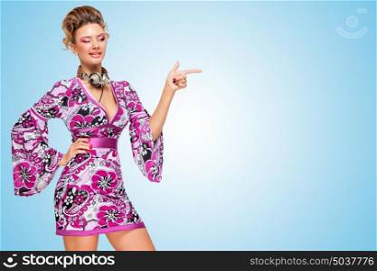 Colorful photo of an astonished fashionable hippie homemaker with metal vintage music headphones around her neck, pointing aside with her finger on blue background.