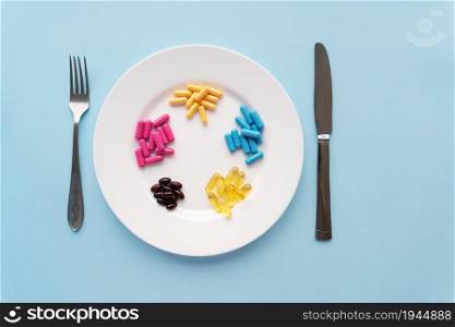 Colorful pills in a large white bowl along with a fork and knife on a blue background. Health concept. Colorful pills in a large white bowl along with a fork and knife on a blue background. Health concept.