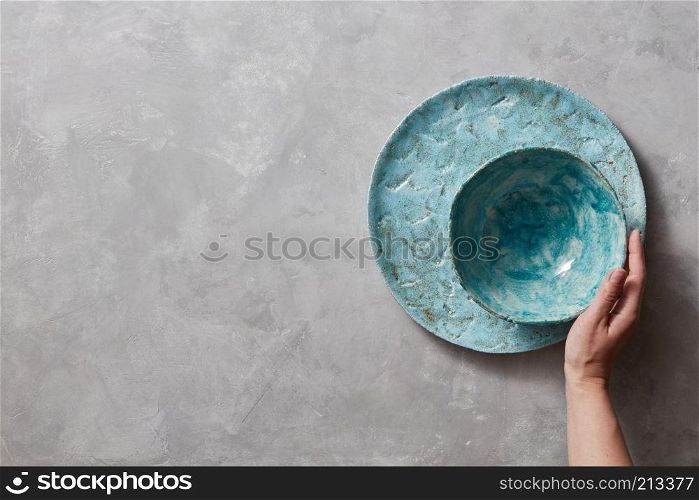 Colorful porceain vintage handmade dishes on a gray marble table with space for text. Woman takes a blue ceramic bowl in her hand . Top view.. Traditional souvenir ceramic handcrafted colorful plate and bowl, holds female hand on a gray concrete table with place under text. Flat lay