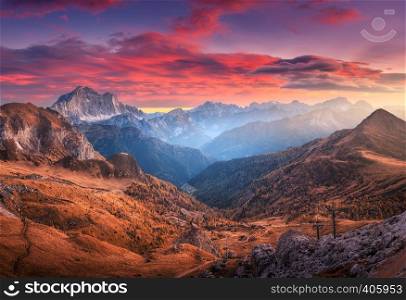 Colorful red sky with clouds over the beautiful mountains in fog at sunset in autumn. Dolomites, Italy. Landscape with mountain range, hills with orange grass, trees, sky with orange sunlight. Travel