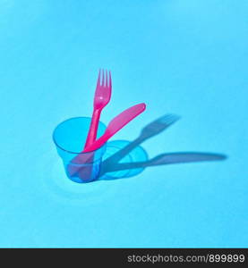 Colorful set of disposable fork and knife in a transparent glass with long shadows, place for text.. Served place with eating plastic utensil and glass with shadows.