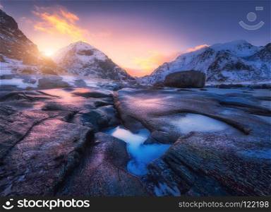 Colorful sunset in Utakleiv beach, Lofoten islands, Norway. Amazing scene with snowy mountains, stones, beautiful reflection in water, red sky, clouds. Winter landscape. Coast with stones in water. Colorful sunset in Utakleiv beach, Lofoten islands, Norway