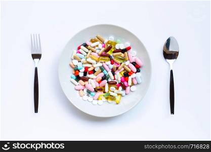 Colorful tablets with capsules and pills on white dish, folk and spoon on white background.