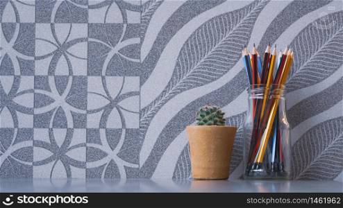 Colorful wooden pencils in glass jar with little green cactus in clay pot on grey tabletop with abstract natural pattern of gray tile wall background