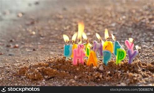 Colourful birthday candles spelling out the alphabet letters - Happy Birthday - standing upright in beach sand burning on a seashore at the edge of the sea