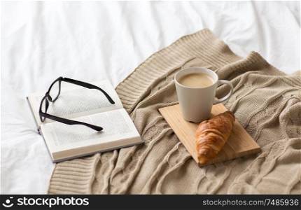 comfort, hygge and interior concept - coffee, croissant, blanket and book on bed at cozy home. coffee, croissant, blanket and book on bed at home