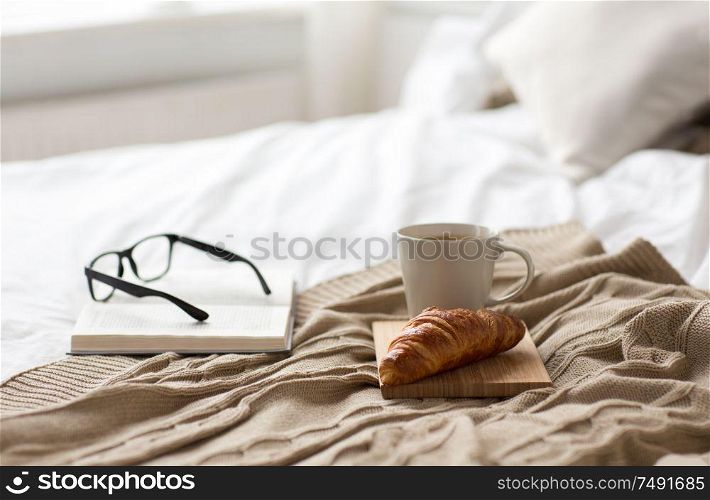 comfort, hygge and interior concept - coffee, croissant, blanket and book on bed at cozy home. coffee, croissant, blanket and book on bed at home