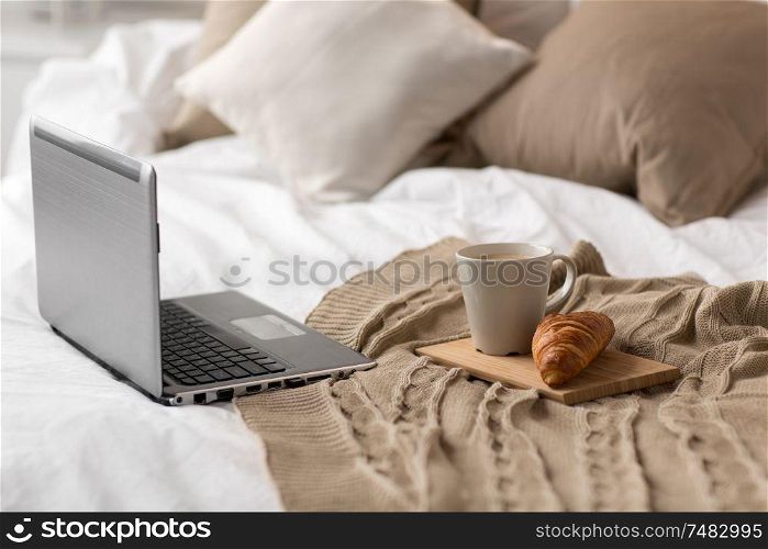 comfort, hygge and interior concept - laptop computer, cup of coffee and croissant on bed at cozy home. laptop, cup of coffee and croissant on bed at home
