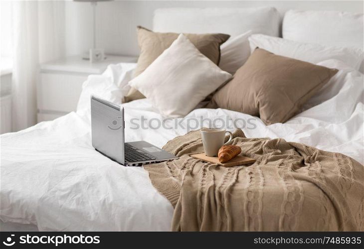 comfort, hygge and interior concept - laptop computer, cup of coffee and croissant on bed at cozy home. laptop, cup of coffee and croissant on bed at home