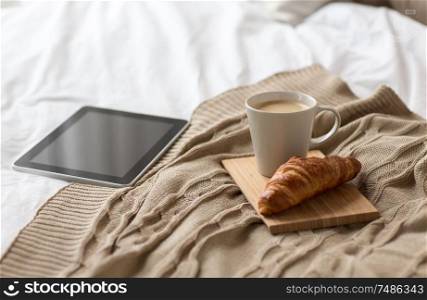 comfort, hygge and interior concept - tablet computer, cup of coffee and croissant on bed at cozy home. tablet pc, coffee cup and croissant on bed at home