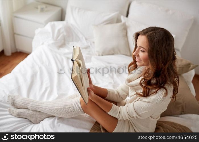 comfort, leisure and people concept - happy young woman reading book in bed at home bedroom