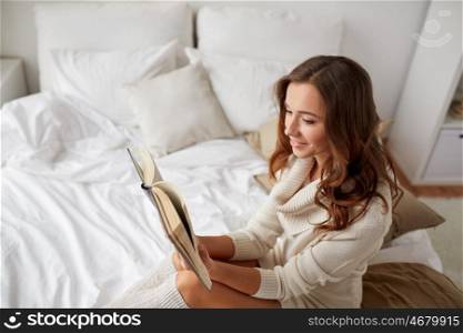 comfort, leisure and people concept - happy young woman reading book in bed at home bedroom