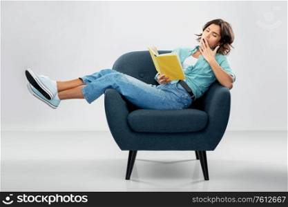 comfort, people and furniture concept - portrait of bored or tired young woman in turquoise shirt and jeans sitting in modern armchair and reading book and yawning over grey background. bored woman in armchair reading book and yawning