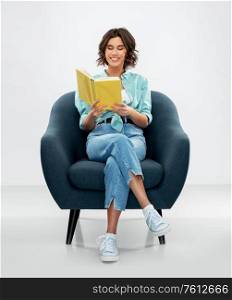 comfort, people and furniture concept - portrait of happy smiling young woman in turquoise shirt and jeans sitting in modern armchair and reading book over grey background. happy young woman in armchair reading book