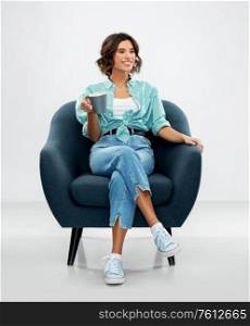 comfort, people and leisure concept - portrait of happy smiling young woman in turquoise shirt and jeans with cup of coffee or tea sitting in modern armchair over grey background. woman sitting in chair with cup of coffee or tea