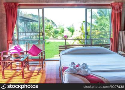 Comfortable tropical style bedroom, sunrise shines through broad windows on glossy wooden floor, green tropical garden at the backyard. Soft focus on the bed.