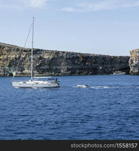 Comino is a small island of the Maltese archipelago between the islands of Malta and Gozo in the Mediterranean Sea. Yacht at the rugged coastline delineated by sheer limestone cliffs, and dotted with deep caves