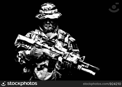 Commando shooter, army special forces rifleman in battle uniform, boonie hat, ammunition in load carrier, camouflaged with paint face, armed assault rifle, half length portrait on black background. Commando soldier in battle ammunition, armed rifle