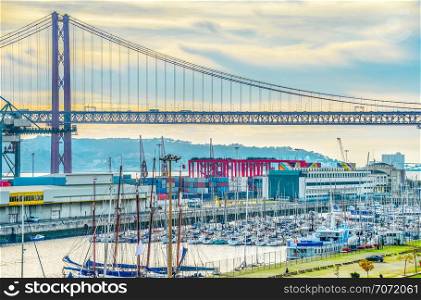 Commercial port of Lisbon and city marina with yachts, 25th April Bridge and Almada skyline under cloudy sky
