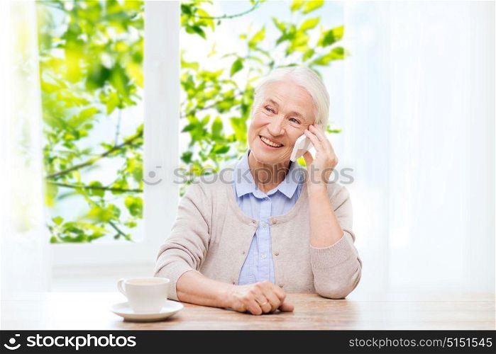 communication, age and people concept - happy senior woman with smartphone and coffee sitting at table and calling at home over window and green natural background. senior woman with smartphone calling at home