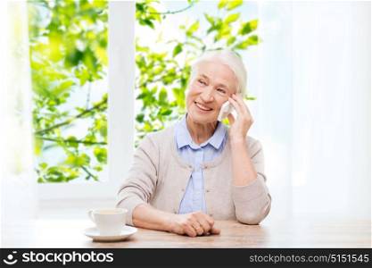communication, age and people concept - happy senior woman with smartphone and coffee sitting at table and calling at home over window and green natural background. senior woman with smartphone calling at home