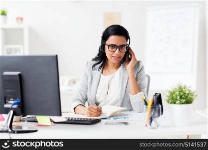 communication, business, people and technology concept - smiling businesswoman or helpline operator with headset and computer talking and writing to notebook at office. businesswoman with headset and notebook at office