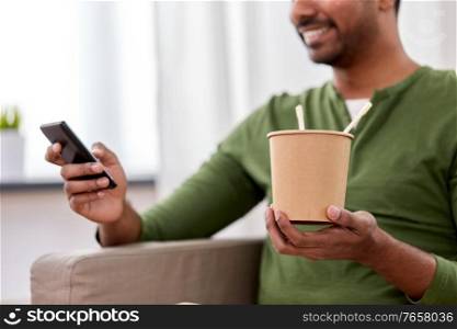 communication, leisure and people concept - smiling indian man with smartphone eating takeaway food at home. indian man with phone eating takeaway food at home