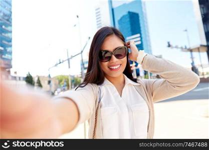 communication, lifestyle and technology concept - smiling young asian woman in sunglasses taking selfie on city street. smiling woman in sunglasses taking selfie in city