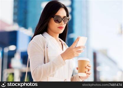 communication, lifestyle and technology concept - young asian woman in sunglasses with takeaway coffee cup and smartphone on city street. woman with smartphone and coffee in city