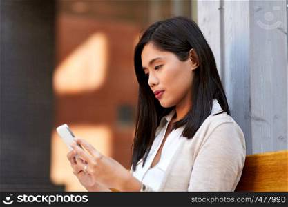 communication, lifestyle and technology concept - young asian woman using smartphone sitting on bench in city yard. asian woman using smartphone sitting on bench