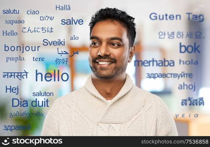 communication, translation and international concept - smiling indian man in knitted woolen sweater at office over greeting words in different foreign languages. indian man over words in foreign languages