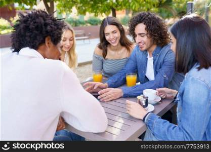 Company of happy multiethnic friends gathering at wooden table in summer cafe and spending time together at weekend. Group of diverse friends chilling in street cafe