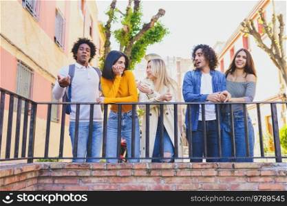 Company of multiethnic friendly people standing near railing in city and talking to each other while spending weekend together. Group of content diverse friends chatting in city