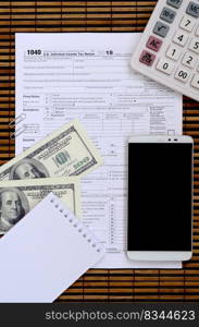 Composition of items lying on the 1040 tax form. Dollar bills, calculator, smartphone, paper clip and notepad. The time to pay taxes