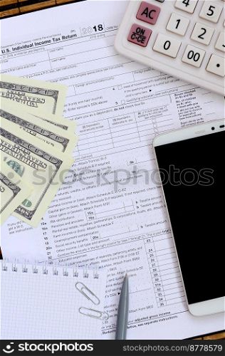 Composition of items lying on the 1040 tax form. Dollar bills, pen, calculator, smartphone, paper clip and notepad. The time to pay taxes