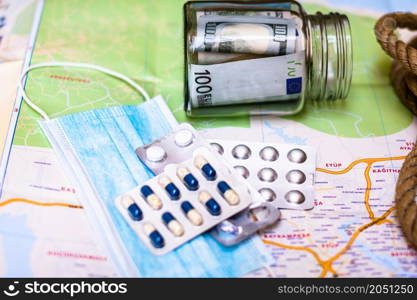 Composition with saving money banknotes in a glass jar with pills and surgical mask on map. Concept of investing and keeping money for healthcare and travel, close up isolated.