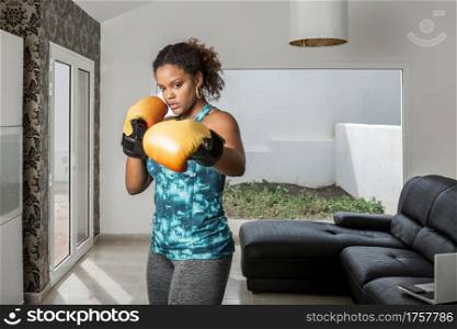Concentrated African American sportswoman in activewear and boxing gloves doing punches while training at home. Confident ethnic woman in boxing gloves during workout at home