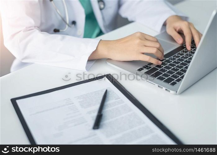 Concentrated Asian woman Doctor hands using laptop computer and working on checklist paper symptoms of patients on a clipboard in the medical room of the hospital.