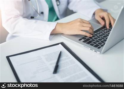 Concentrated Asian woman Doctor hands using laptop computer and working on checklist paper symptoms of patients on a clipboard in the medical room of the hospital.