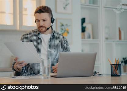 Concentrated bearded man freelancer or entrepreneur wearing headset holding document and looking at it during online call while working at home and using laptop. Remote work and freelance. Young man analyzing information during an online call at home