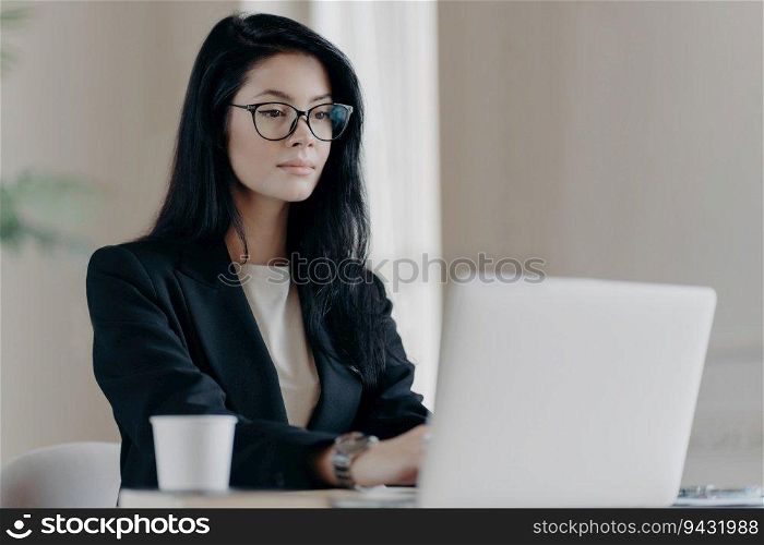 Concentrated brunette manager in coworking space, wearing glasses, creates website post on laptop with coffee.