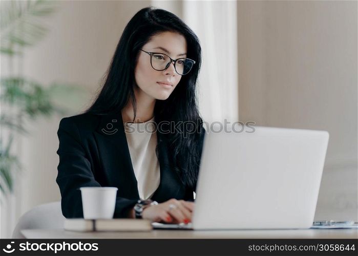 Concentrated brunette woman IT specialist in eyewear searches information using internet, keyboards on laptop computer, drinks coffee, has serious expression. Business and freelance concept.