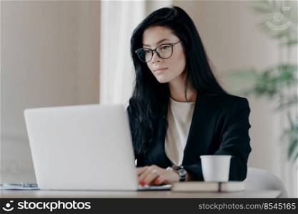Concentrated brunette woman IT specialist in eyewear searches information using internet, keyboards on laptop computer, drinks coffee, has serious expression. Business and freelance concept.