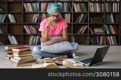 Concentrated college female student sitting on the floor, surronded by heap of books, researching with and laptop in university library. Serious hipster girl with blue hair and trendy eyewear studying in library with laptop on the internet.